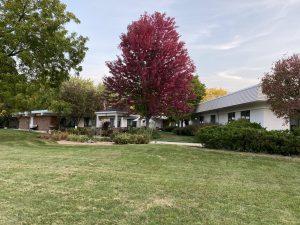 An exterior shot of a senior living facility. The facility is white with a black roof and is surrounded by multicolored trees and lush lawns.