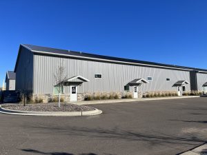 An exterior shot of a gray building. The building has a black roof and many well-kept plants lining the exterior.